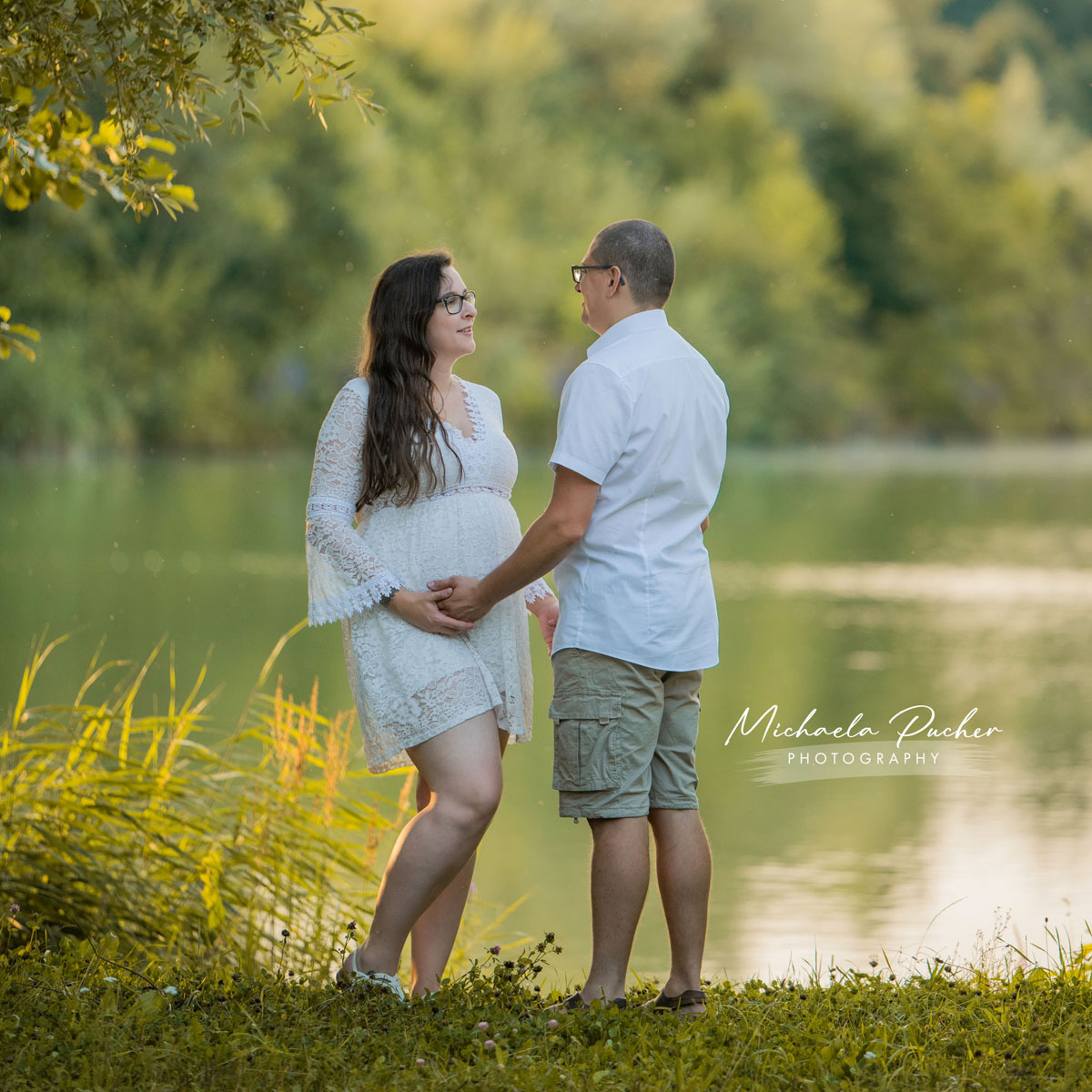 Schwangere Frau und ihr Partner stehen am See im warmen Abendlicht, halten gemeinsam den Babybauch und blicken sich liebevoll an.